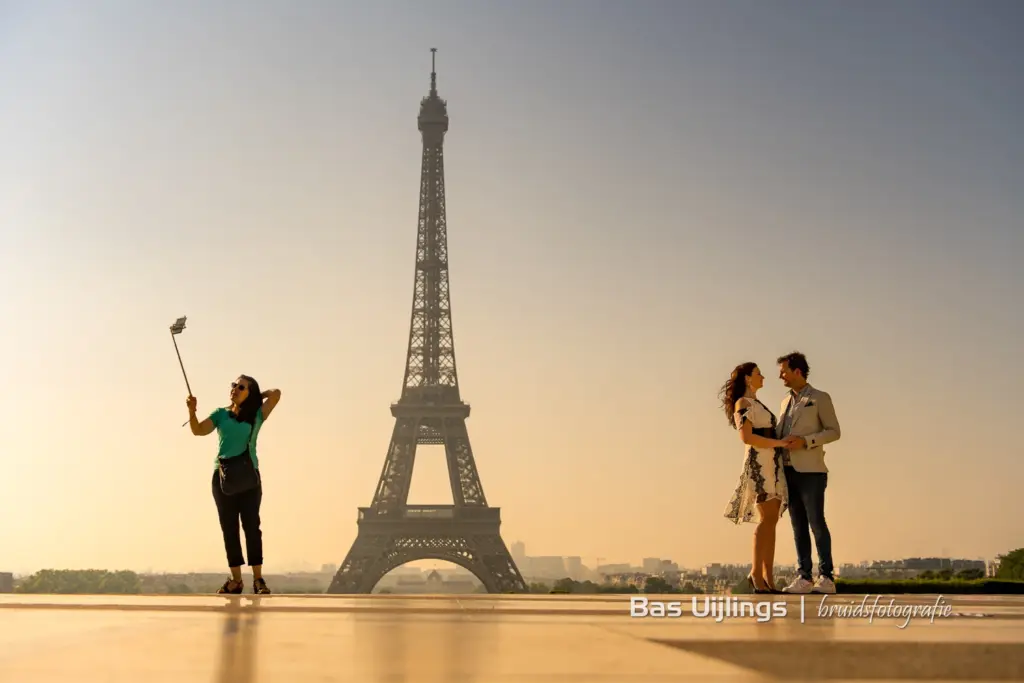 een foto van een man en een vrouw rechts in de foto die elkaar vast houden, de Eiffel toren op de achtergrond en links staan een vrouw met een selfie stick die een foto maakt