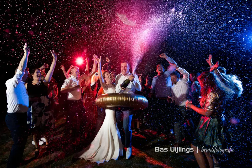 een bruid en bruidegom die in een opblaasband staan, de bruidegom spuit met champagne en om hun heen staan vrienden en familie met hun handen in de lucht. blauwe en roze belichtingen