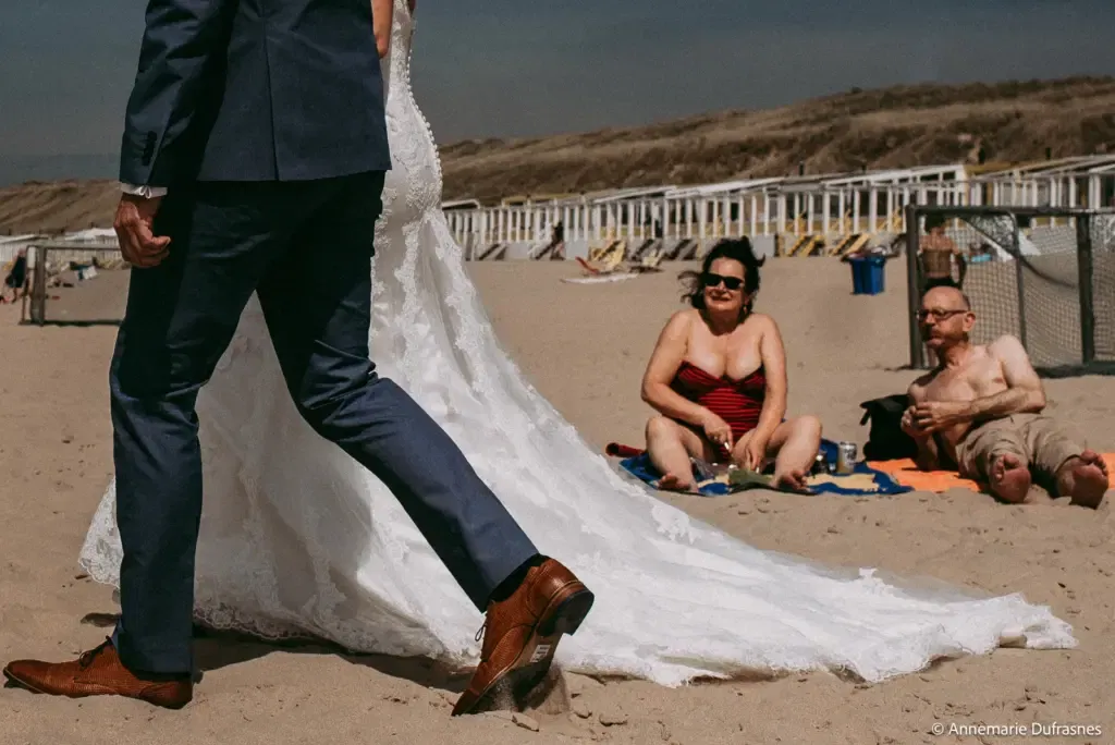 een bruidspaar die over het straks loopt en worden aangekeken door een man en een vrouw die op het strand zitten