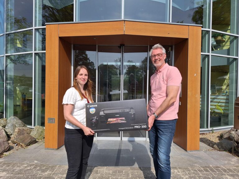 Laura Bonne en Remco Rutgers staan glimlachend buiten een modern glazen kantoorgebouw, waarbij Laura Bonne een grote doos met een Canon printer vasthoudt.
