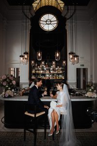 Een pasgetrouwd stel geniet van een romantisch moment aan een stijlvolle bar met een marmeren blad, omringd door sfeervolle verlichting en luxe interieurdetails. De bruidegom draagt een elegant donker pak, terwijl de bruid straalt in een moderne witte jurk met een lange sluier. Ze proosten met champagne, terwijl bloemen en gedempt licht een intieme sfeer creëren in deze chique, historische locatie.