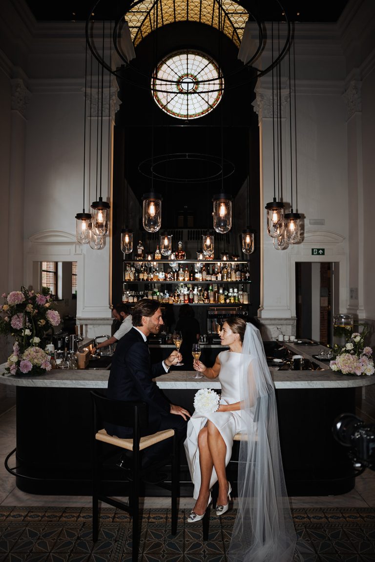 Een pasgetrouwd stel geniet van een romantisch moment aan een stijlvolle bar met een marmeren blad, omringd door sfeervolle verlichting en luxe interieurdetails. De bruidegom draagt een elegant donker pak, terwijl de bruid straalt in een moderne witte jurk met een lange sluier. Ze proosten met champagne, terwijl bloemen en gedempt licht een intieme sfeer creëren in deze chique, historische locatie.