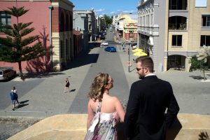 Een stijlvol gekleed stel tijdens hun trouwerij staat bovenaan een trap en kijkt uit over een zonnige, historische straat in Perth, Australië. De vrouw draagt een lichte, bloemige jurk en de man een zwart pak met een zonnebril. Op de achtergrond zijn klassieke gebouwen, voorbijgangers en geparkeerde auto's te zien, wat een levendige stedelijke sfeer creëert.