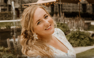 Marilyn Hansum lacht naar de camera in een zomerse tuin met fontein en sfeerverlichting op de achtergrond.