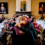 Emotioneel moment op een bruiloftsfeest waar drie vrouwen elkaar omhelzen terwijl gasten aan lange tafels applaudisseren.