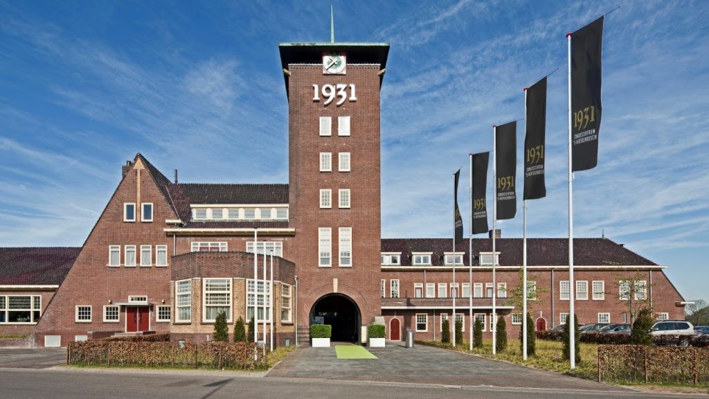 Vooraanzicht van evenementenlocatie 1931 Congrescentrum in ’s-Hertogenbosch, waar de MPN Fotovakbeurs wordt gehouden; karakteristiek bakstenen gebouw met hoge centrale toren, klok en vlaggen met het logo ‘1931’ onder een blauwe lucht.
