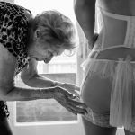 Oudere vrouw helpt bruid met haar lingerie tijdens de voorbereidingen op de trouwdag, intiem en eerlijk gefotografeerd door Isabelle Hattink.