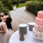 Bruiloftstaferel met kind in feestjurk dat uit een leeg glas drinkt naast een grote witte bruidstaart met roze bloemen.