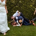 Speels bruiloftsmoment waarbij een groep mannen lachend op elkaar valt op het gras terwijl gasten eromheen lopen.