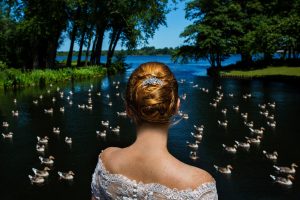 Vrouw in trouwjurk kijkt uit over een rustige vijver vol eenden, omringd door groene bomen op een zonnige dag – een symbolisch beeld dat de vraag ‘Wat is echt zien?’ oproept.