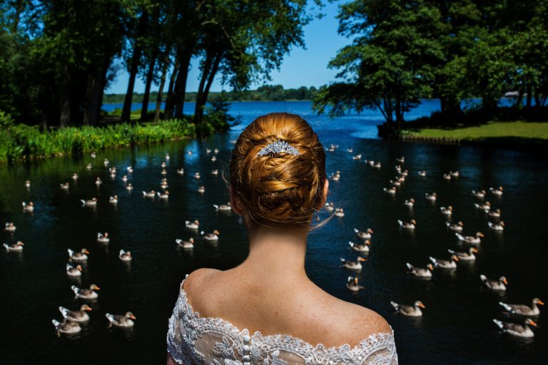 Vrouw in trouwjurk kijkt uit over een rustige vijver vol eenden, omringd door groene bomen op een zonnige dag – een symbolisch beeld dat de vraag ‘Wat is echt zien?’ oproept.